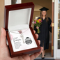 Graduation Cap Name Necklace displayed in wooden gift box with a blurred graduate holding flowers in the background