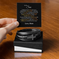 Men's cross bracelet for son made of black leather and metal displayed with a love note from mom